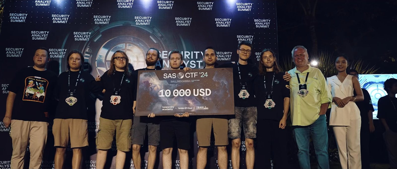 The winning team holding a 10,000 USD prize board on stage at SAS CTF 2024 in Bali